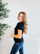 Woman wearing a black top and blue jeans standing against a white wall with a plant to the left.
