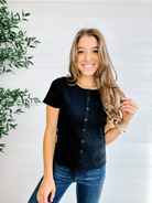 Woman wearing a black button-up shirt and blue jeans standing against a white brick wall with a plant to the left.