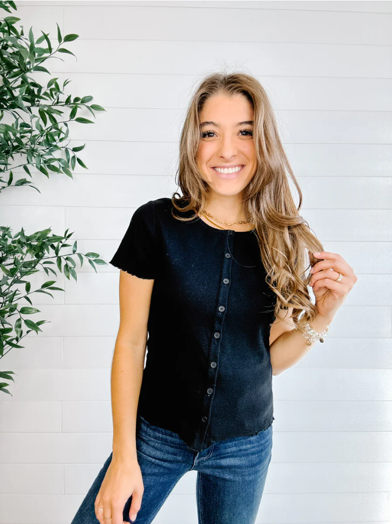 Woman wearing a black button-up shirt and blue jeans standing against a white brick wall with a plant to the left.