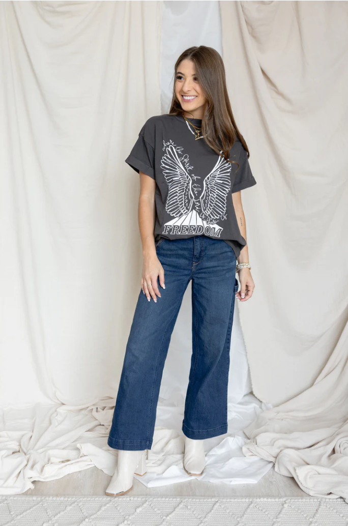 Woman wearing a graphic t-shirt and blue jeans standing against a white curtain.