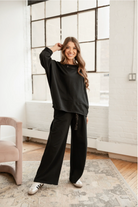 Woman in black outfit standing in a room with large windows and a white wall.