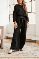 Woman wearing a black outfit with wide-leg pants in a room with large windows.