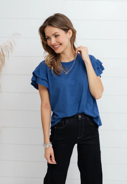 Woman wearing a blue ruffled blouse and black pants against a white wooden panel background