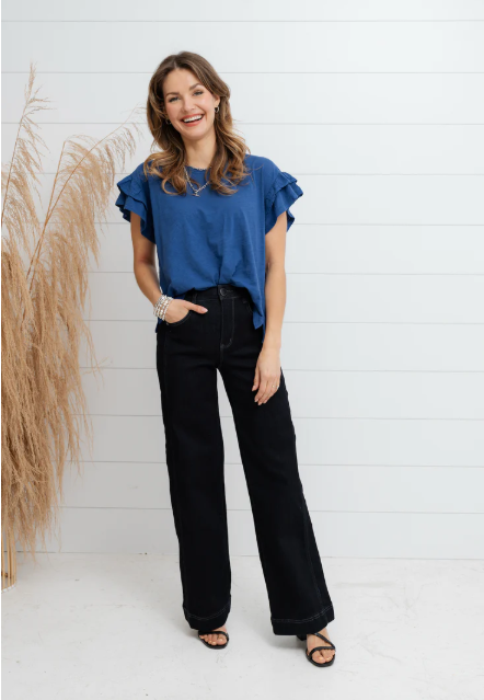 Woman wearing a blue top and black pants standing against a white wall with decorative elements.