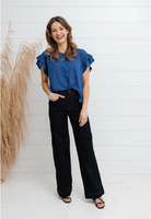 Woman wearing a blue top and black pants standing against a white wall with decorative elements.