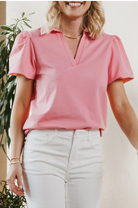Woman wearing a pink blouse with puffed sleeves and white pants, standing indoors.