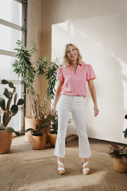 Woman in a pink shirt and white pants standing in a room with plants.