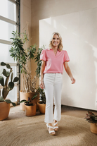 Woman in a pink shirt and white pants standing in a room with plants.