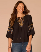 A woman wearing a black bohemian-style blouse with gold, blue, and white floral embroidery on the neckline and sleeves, paired with blue jeans, set against a solid beige background.