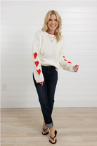 Woman wearing a white sweater with red heart patterns and dark jeans against a white wall.
