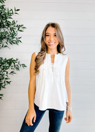 Woman wearing a white sleeveless top and blue jeans standing against a white wall with a plant to the left.