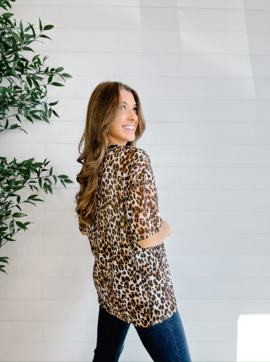 Woman wearing a leopard print shirt standing next to a plant against a white wall
