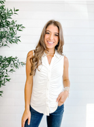 Woman wearing a white sleeveless top and blue jeans standing against a white wall with a plant to the left.