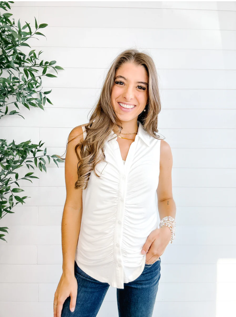 Woman wearing a white sleeveless top and blue jeans standing against a white wall with a plant to the left.