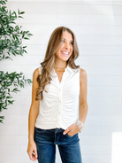 Woman wearing a white sleeveless vest and blue jeans standing against a white wall with a plant to the left.