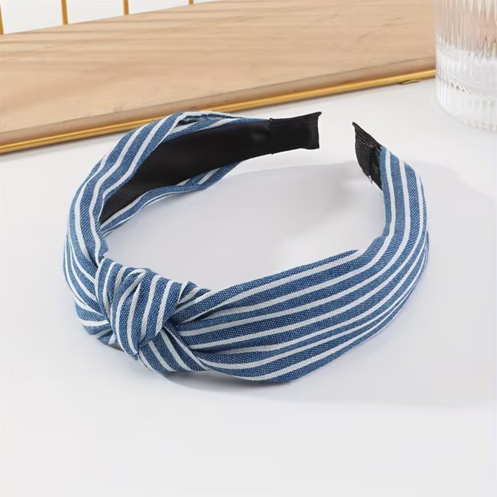 A wide fabric headband with a twisted knot detail, featuring blue and white vertical stripes. The headband is displayed on a white surface with a wooden tray and a glass in the background.