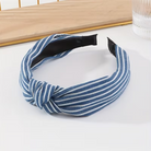 A wide fabric headband with a twisted knot detail, featuring blue and white vertical stripes. The headband is displayed on a white surface with a wooden tray and a glass in the background.