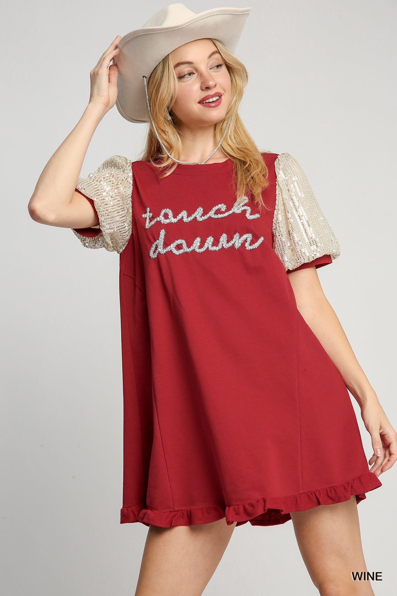 Woman wearing a red dress with 'touch dawn' text and sequin sleeves, holding a beige hat.