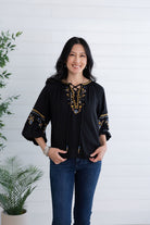A woman wearing a black long-sleeve blouse with gold and blue embroidered details on the neckline and cuffs, paired with dark blue jeans, standing against a white shiplap wall with potted plants.