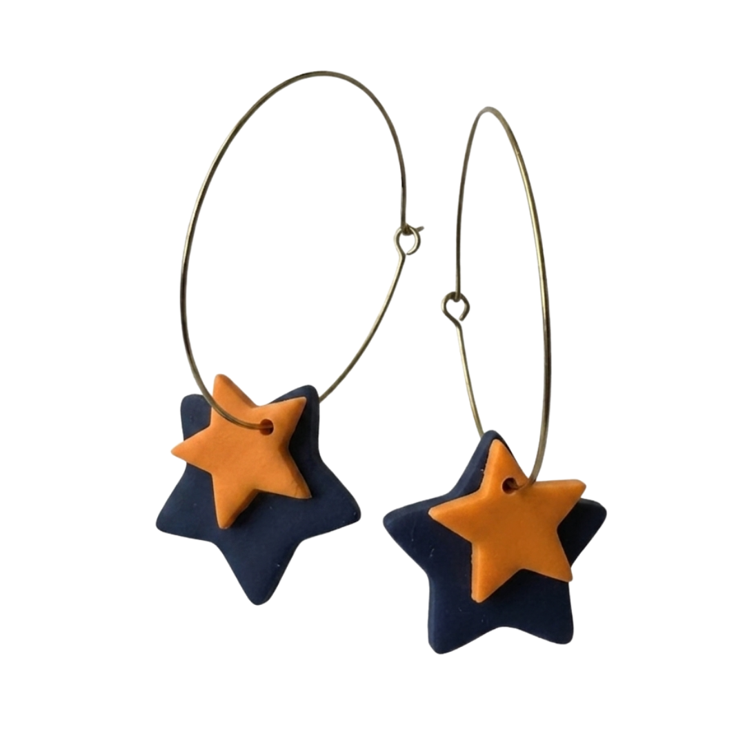 A pair of gold hoop earrings with dangling star-shaped charms, featuring a dark navy blue star base and a smaller orange star layered on top, shown against a plain white background.