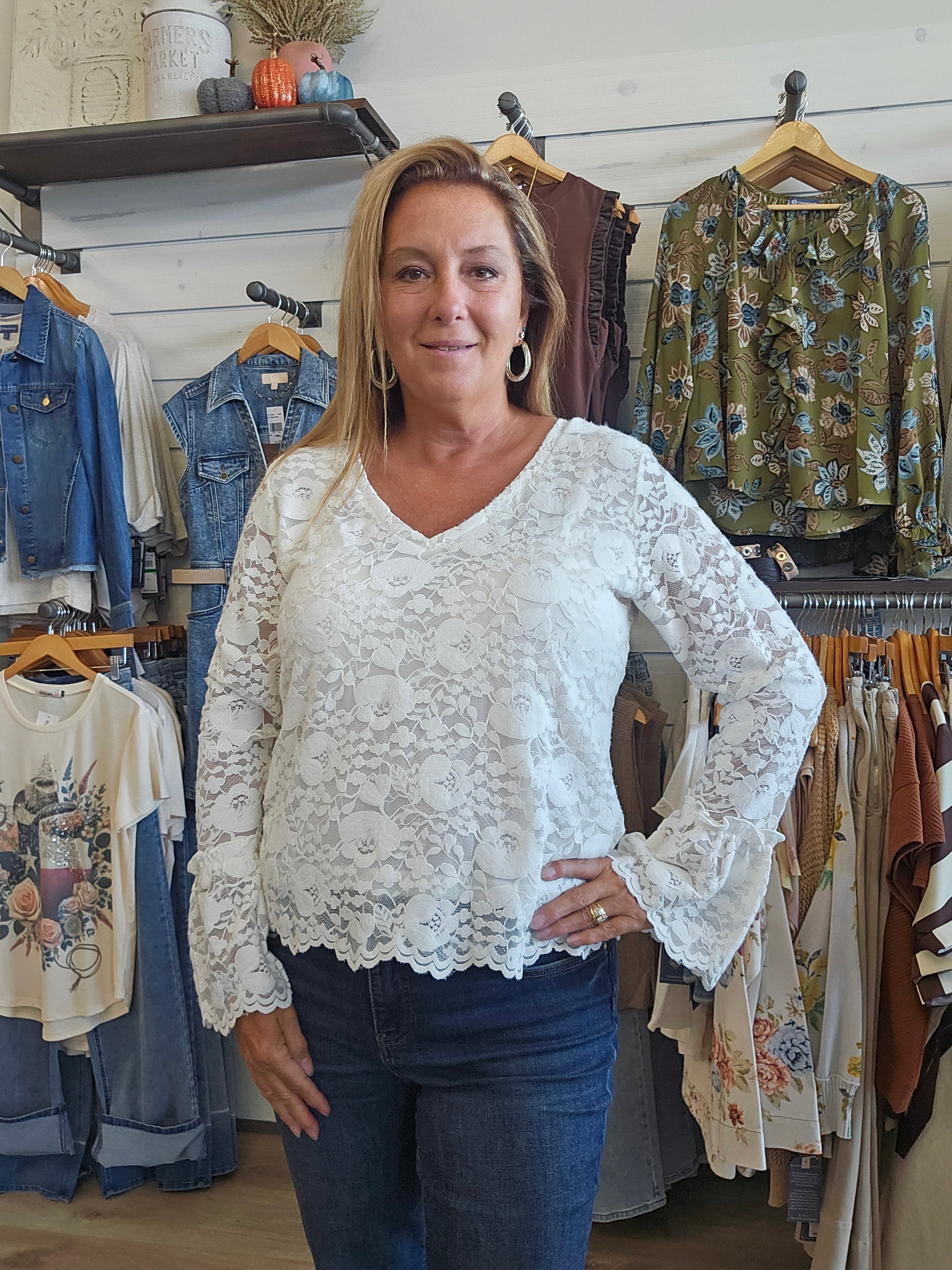 A woman with blonde hair stands in a clothing boutique wearing a white lace long-sleeve blouse with a V-neck and blue jeans. Clothing racks with various garments are visible in the background.