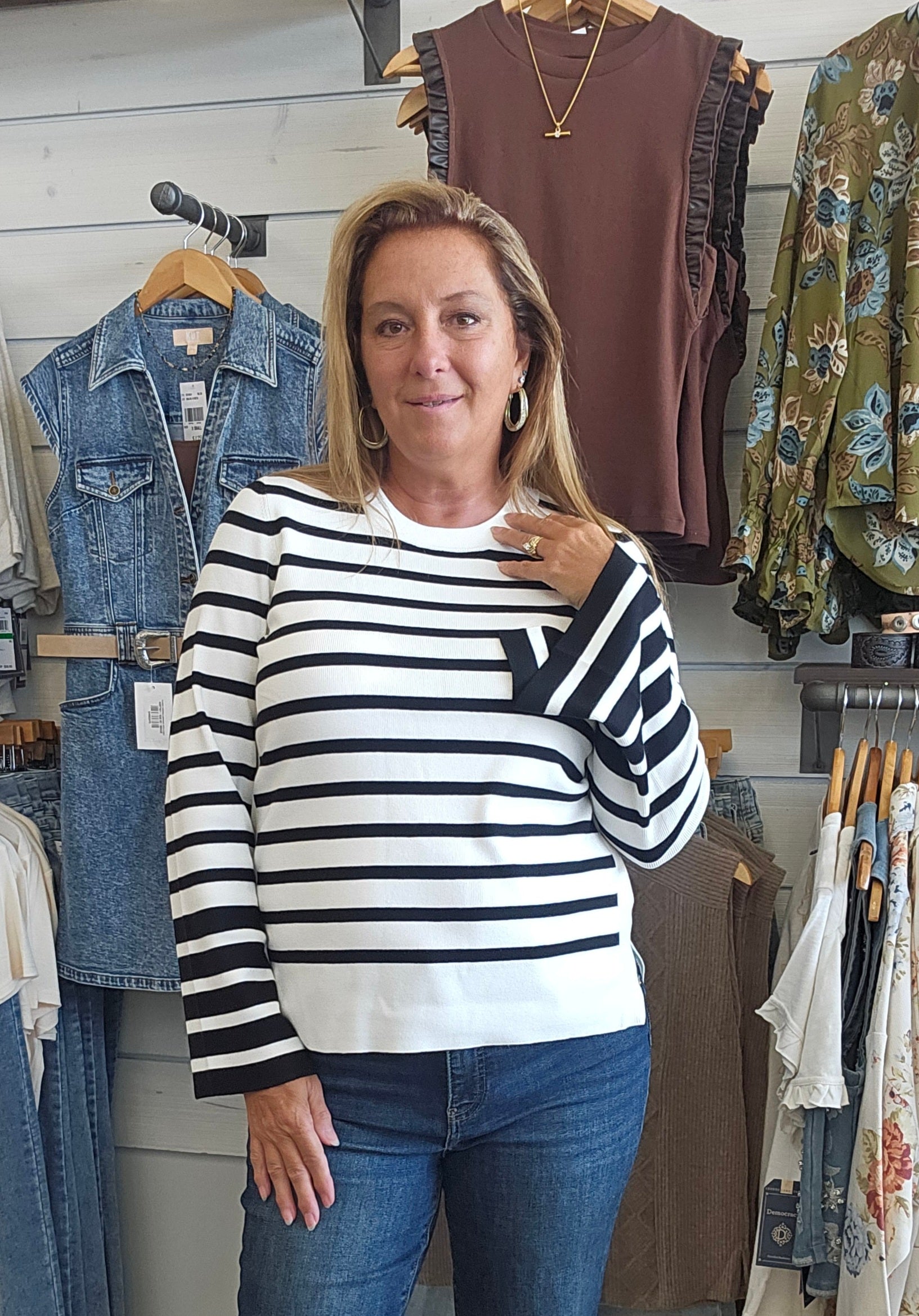 A woman with blonde hair stands in a clothing store wearing a black and white horizontally striped long-sleeve top and blue jeans. Clothing racks with various garments are visible in the background.