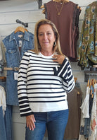 A woman with blonde hair stands in a clothing store wearing a black and white horizontally striped long-sleeve top and blue jeans. Clothing racks with various garments are visible in the background.
