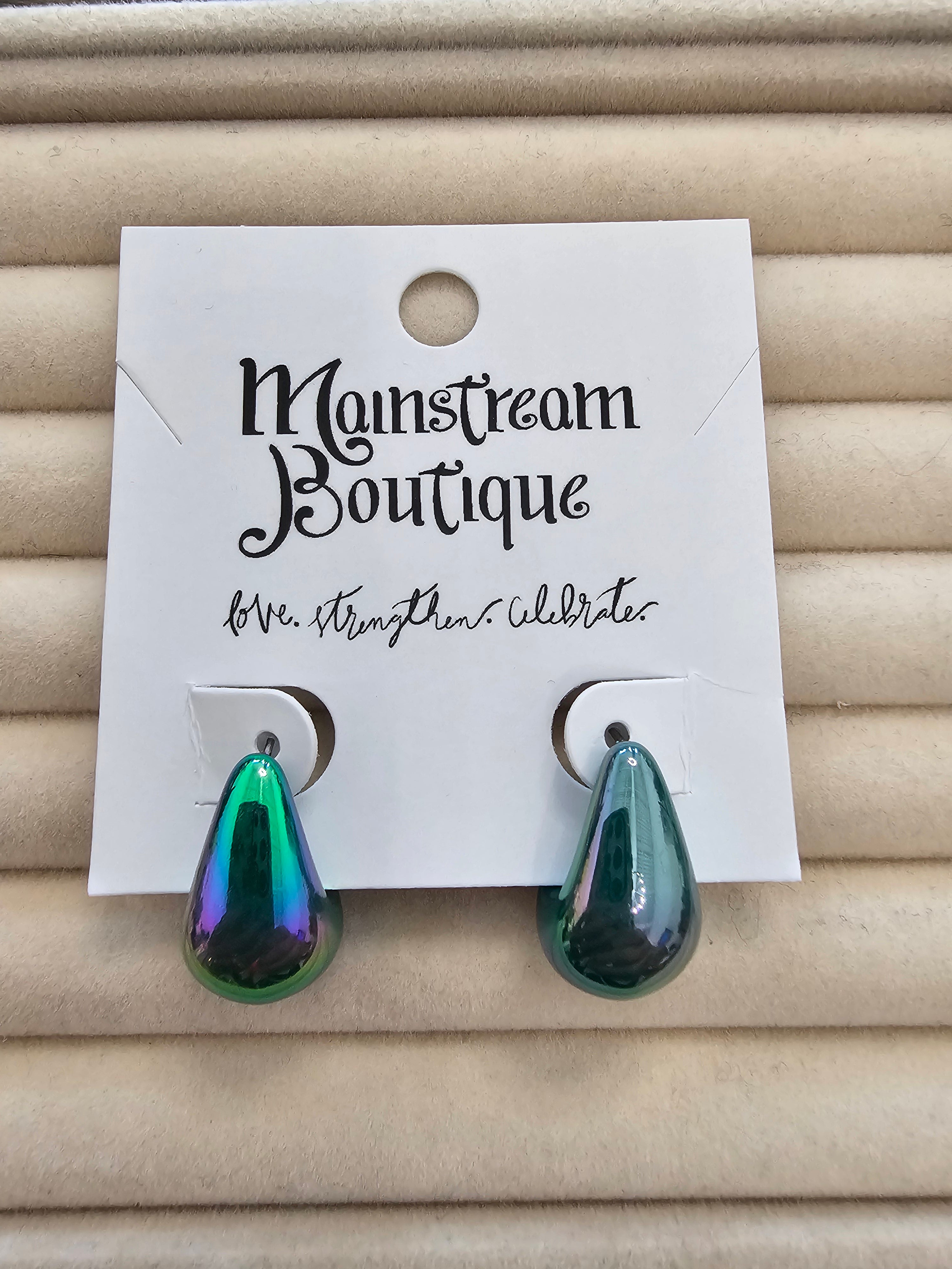A pair of teardrop-shaped drop earrings with an iridescent green and blue finish, displayed on a white card labeled 'Monstream Boutique' against a beige textured background.