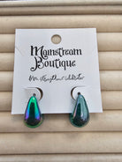 A pair of teardrop-shaped drop earrings with an iridescent green and blue finish, displayed on a white card labeled 'Monstream Boutique' against a beige textured background.
