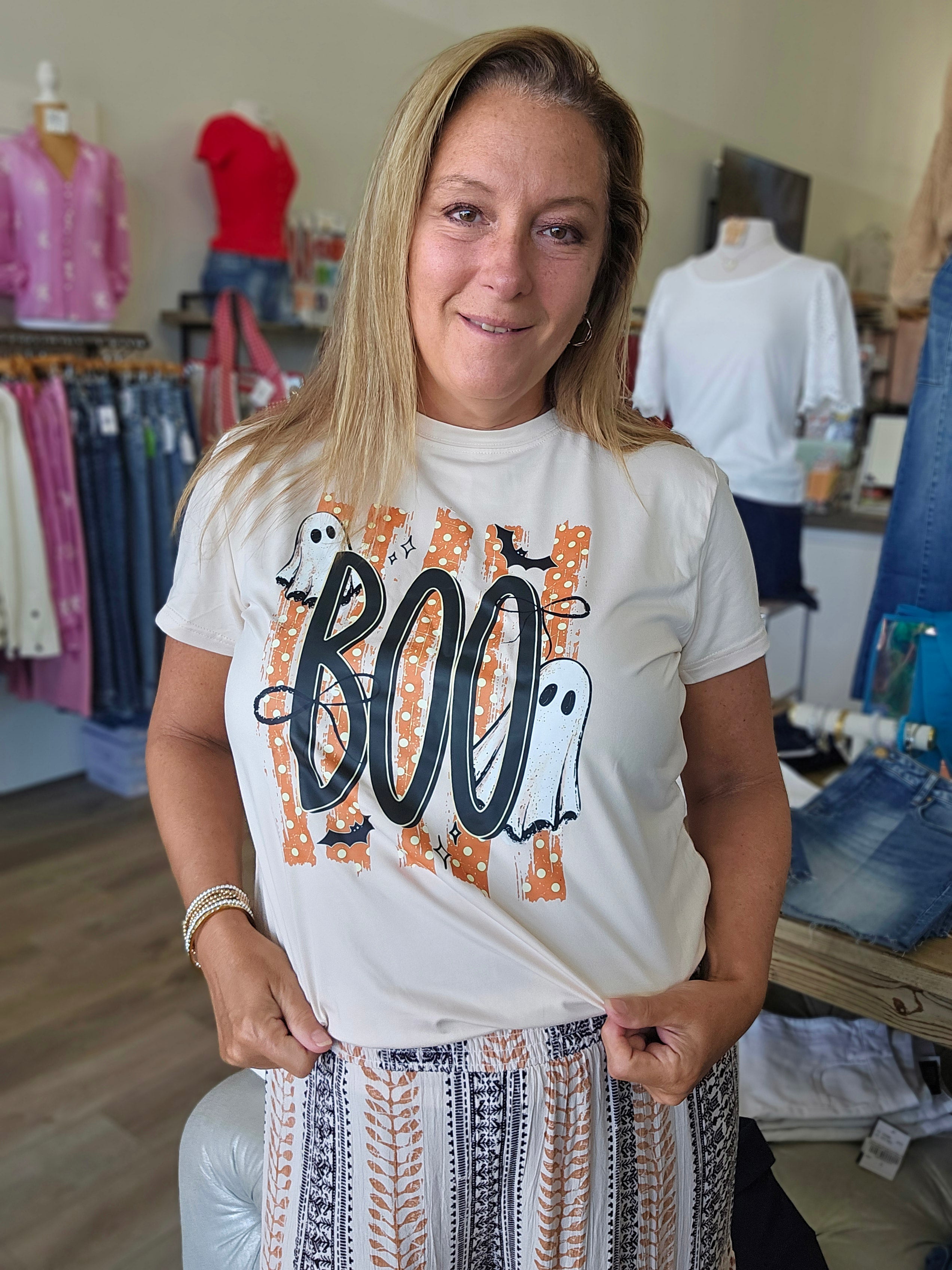 A woman wearing a white graphic t-shirt with a Halloween 'BOO' design in black and orange, paired with a patterned skirt, standing inside a clothing store.