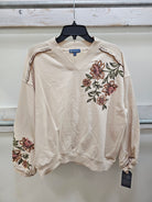 A cream-colored long-sleeve V-neck sweatshirt with pink and green floral embroidery on the chest and sleeve, hanging on a black hanger against a light gray wall.