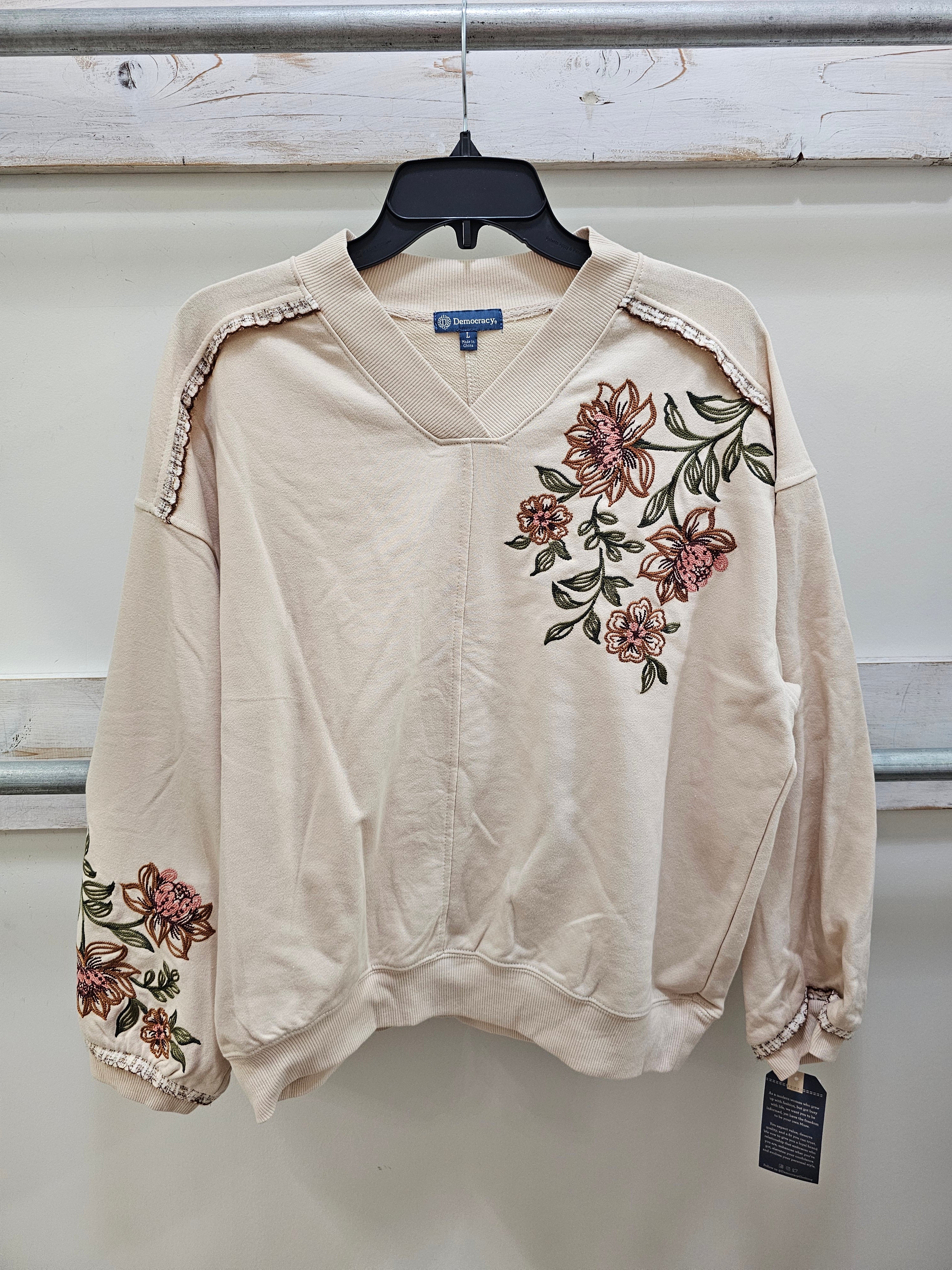 A cream-colored long-sleeve V-neck sweatshirt with pink and green floral embroidery on the chest and sleeve, hanging on a black hanger against a light gray wall.