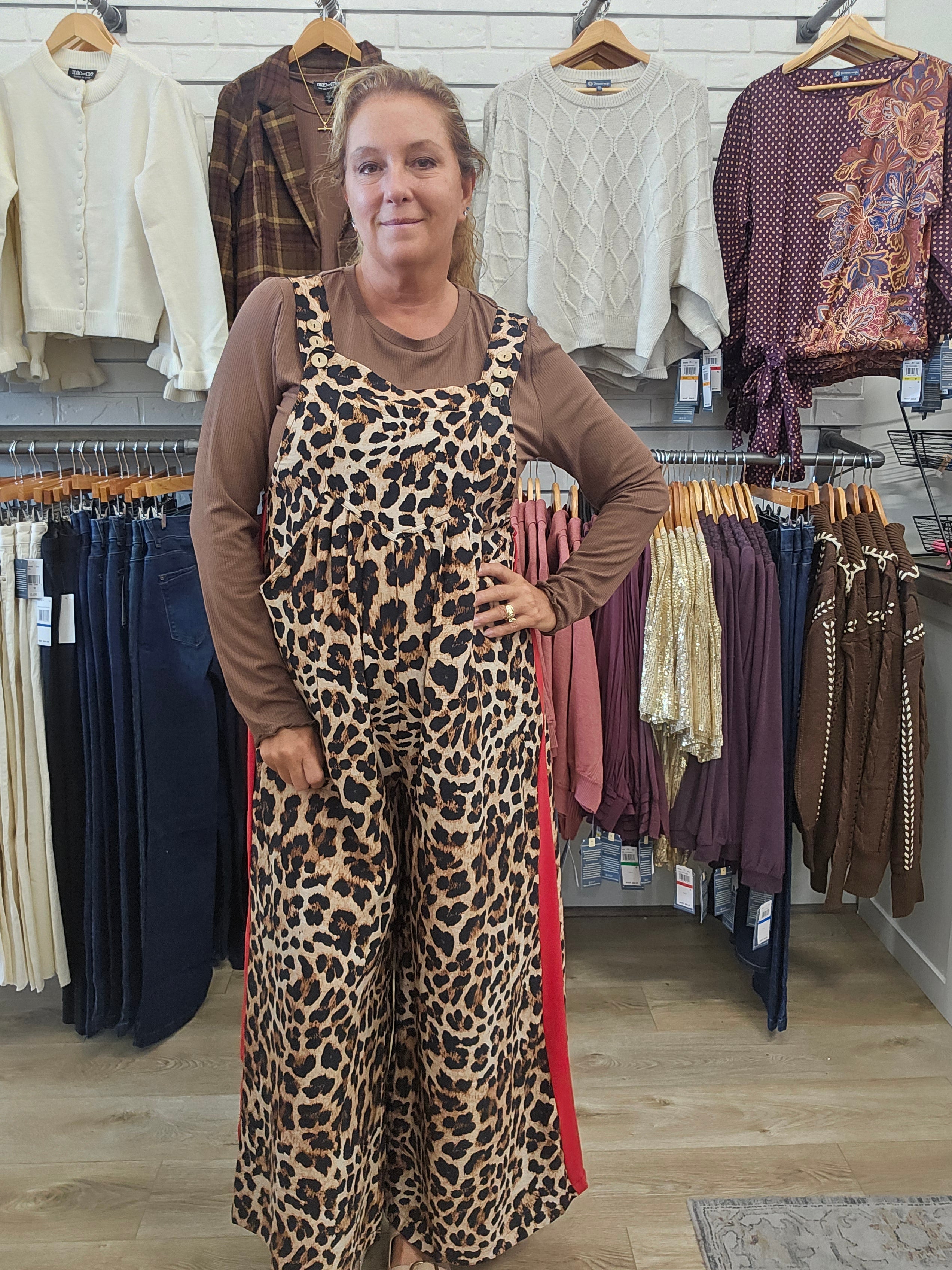A woman modeling a leopard print overall jumpsuit with red side stripes over a brown long-sleeve shirt, standing inside a clothing store.