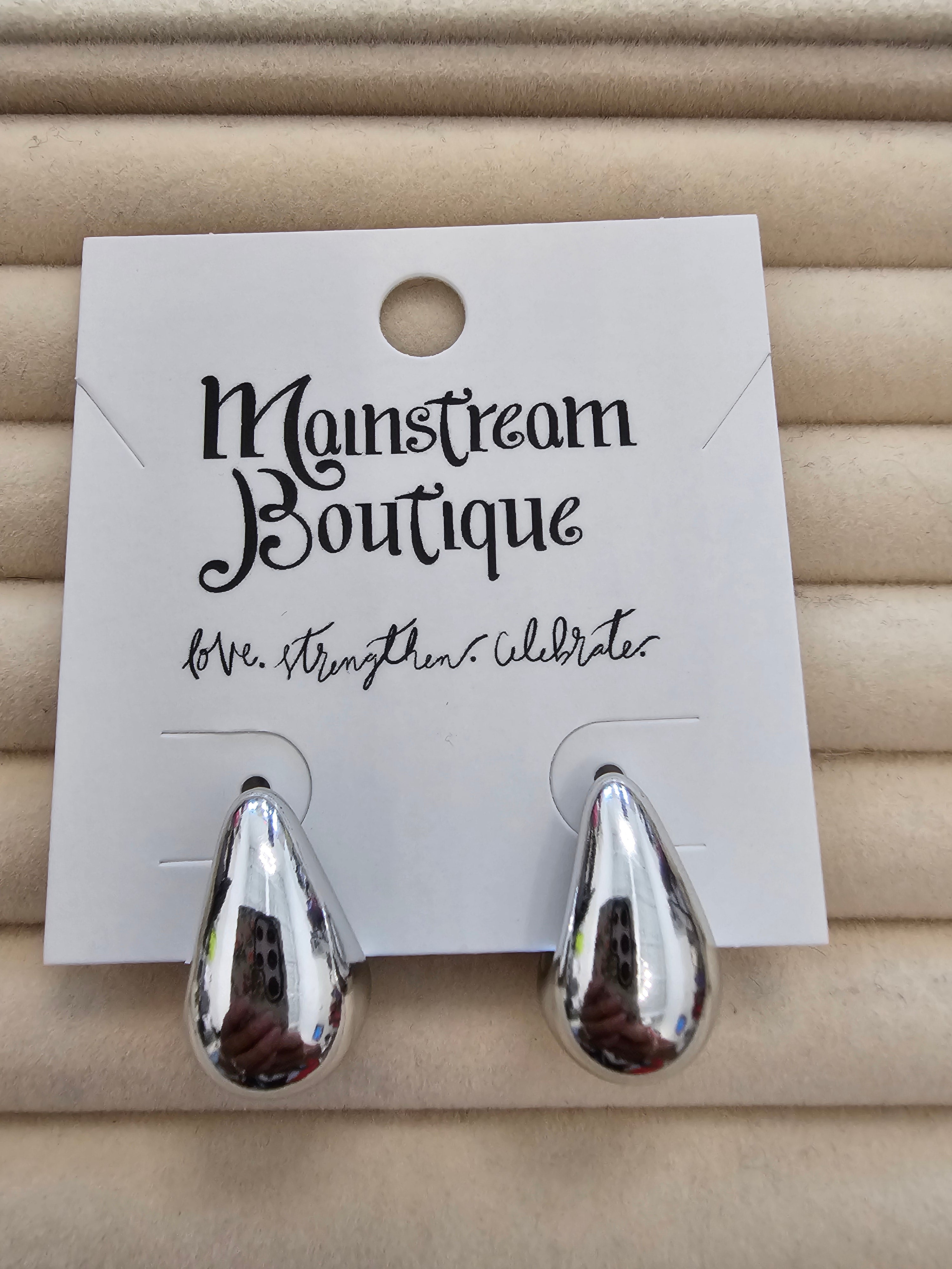 A pair of shiny silver teardrop-shaped earrings displayed on a white card labeled 'Monstream Boutique', set against a beige textured background.