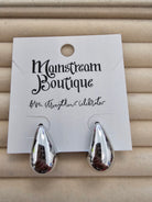 A pair of shiny silver teardrop-shaped earrings displayed on a white card labeled 'Monstream Boutique', set against a beige textured background.
