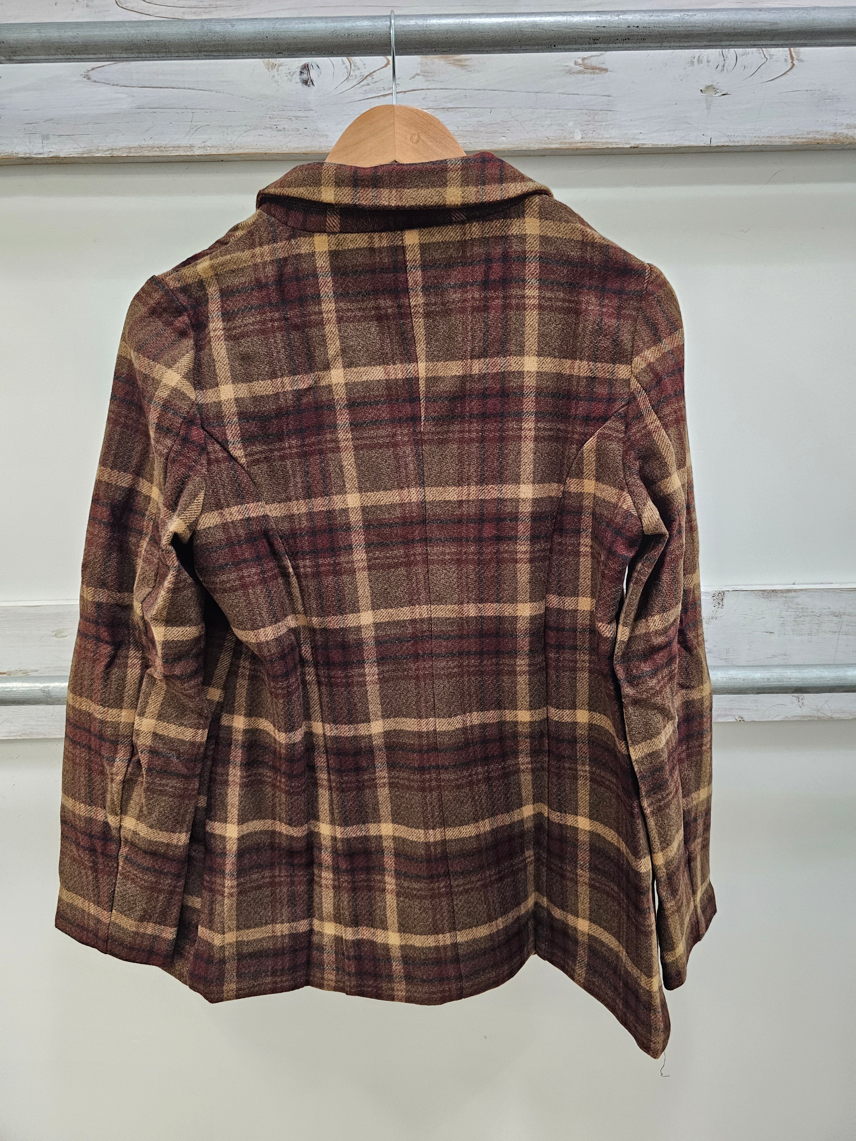 A vintage plaid flannel shirt jacket hanging on a wooden hanger against a white wall with rustic wooden beams.