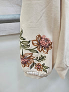 Close-up of a cream-colored sweatshirt sleeve featuring embroidered pink and brown flowers with green leaves, finished with a frayed ribbed cuff.