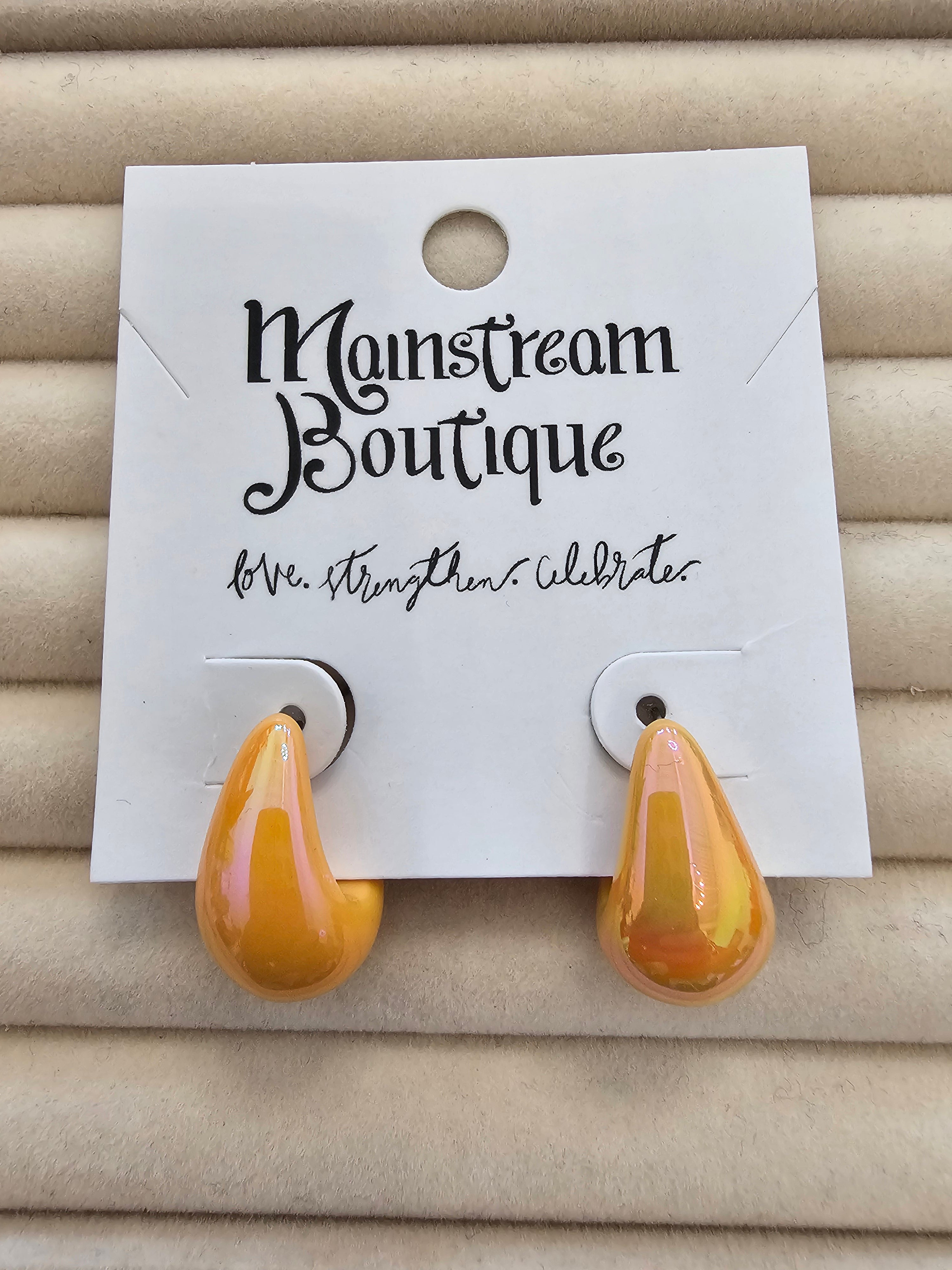 A pair of glossy amber-yellow teardrop-shaped earrings displayed on a white backing card labeled 'Monstream Boutique', set against a beige textured background.