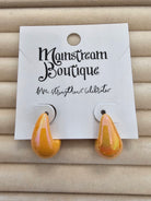 A pair of glossy amber-yellow teardrop-shaped earrings displayed on a white backing card labeled 'Monstream Boutique', set against a beige textured background.