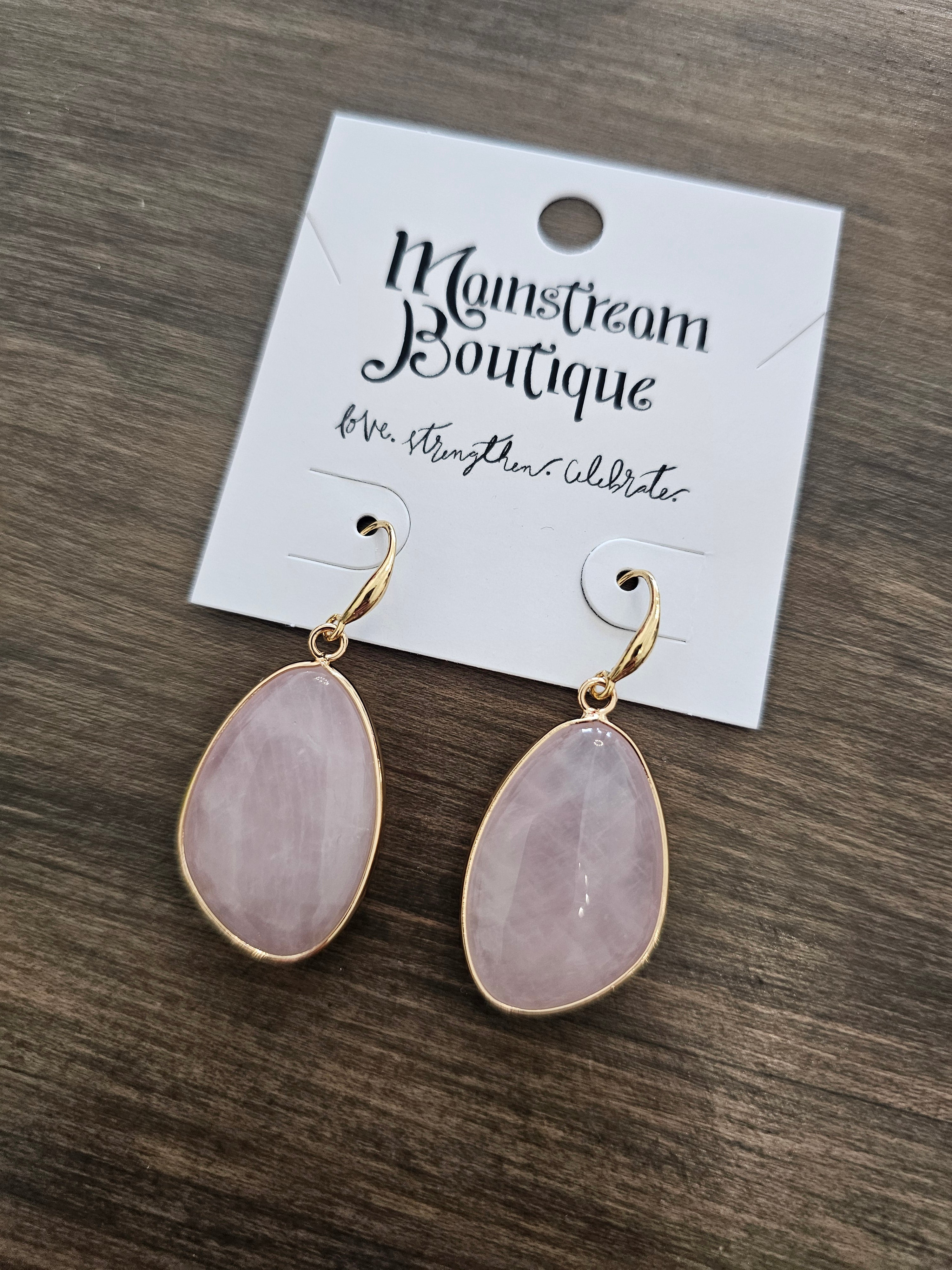 A pair of teardrop-shaped rose quartz drop earrings with gold-plated settings, attached to a white display card labeled 'Monsieur Boutique', placed on a dark wood surface.