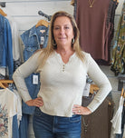 A woman modeling a cream-colored, long-sleeve ribbed henley top with three buttons, paired with blue jeans, standing in a clothing boutique.