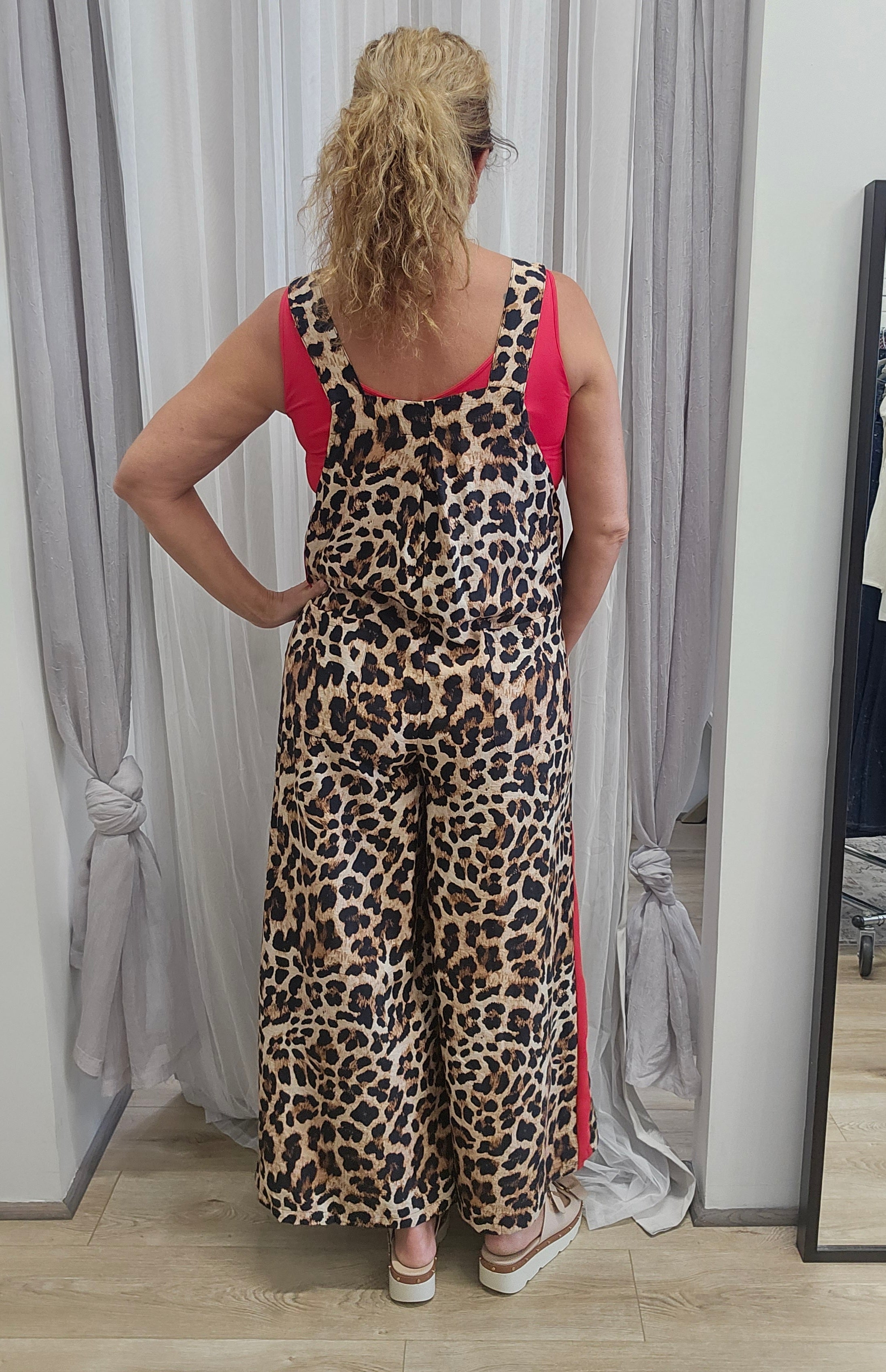 A woman stands with her back to the camera wearing a leopard-print wide-leg jumpsuit with bright pink side panels and straps, in front of gray curtains.
