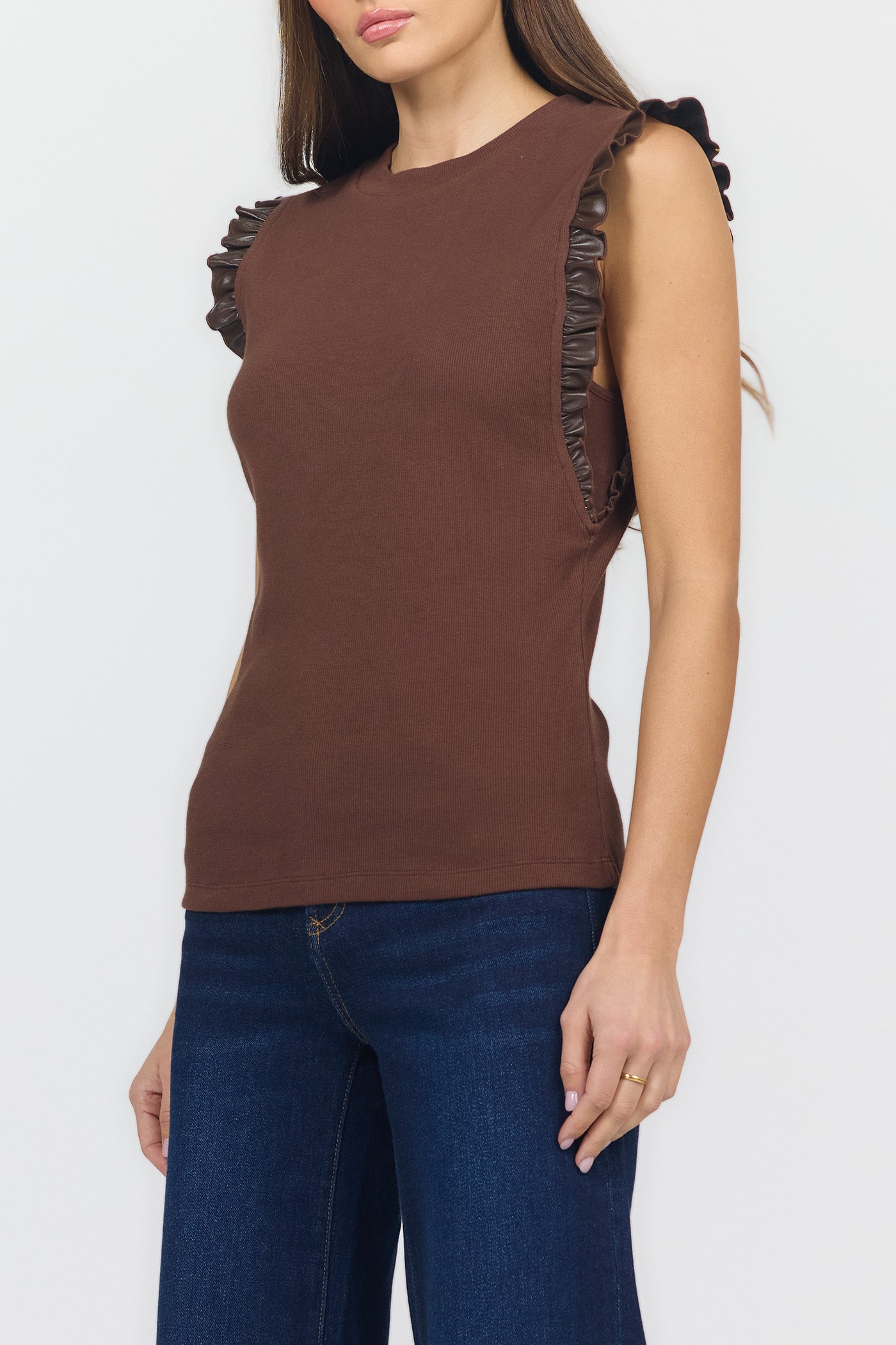 A woman wearing a sleeveless brown knit top with dark ruffled trim along the shoulders and blue jeans, photographed against a light gray studio background.