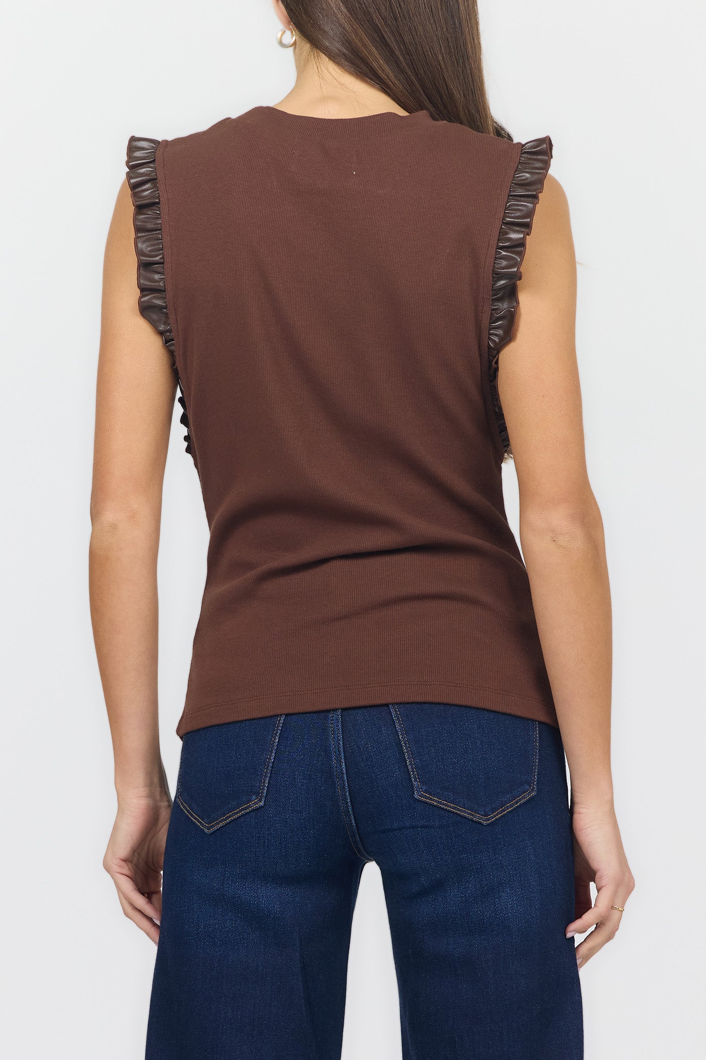 A woman shown from the back wearing a sleeveless brown top with dark ruffled trim along the shoulders and dark blue jeans against a light gray studio background.