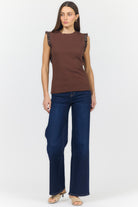 A woman modeling a sleeveless brown top with ruffled shoulder details and dark blue wide-leg jeans against a light gray studio background.
