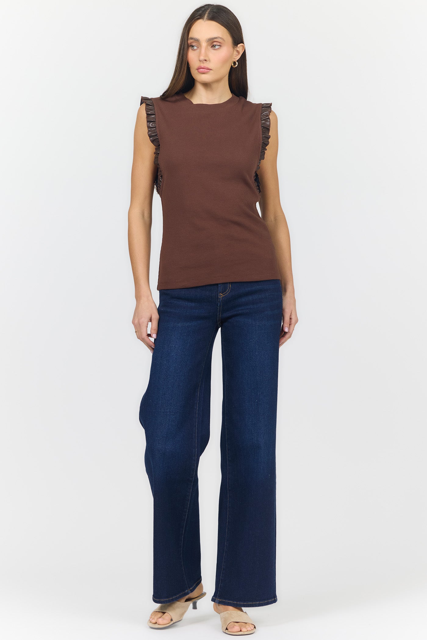 A woman modeling a sleeveless brown top with ruffled shoulder details and dark blue wide-leg jeans against a light gray studio background.