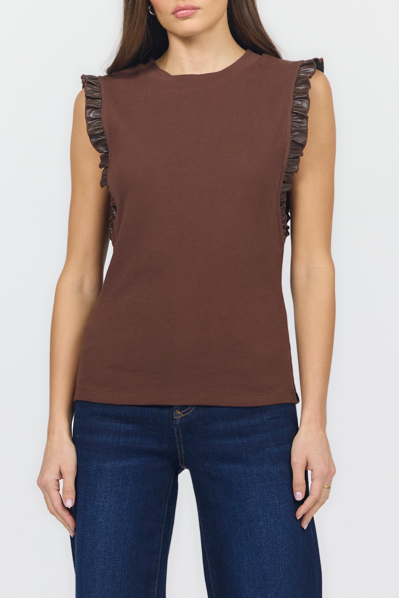 A woman wearing a sleeveless brown top with dark ruffled trim along the shoulders, paired with dark blue jeans, against a light gray studio background.